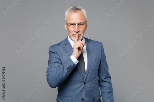 Secret elderly gray-haired bearded business man in blue suit shirt tie isolated on grey background . Achievement career wealth business concept. Saying hush be quiet with finger on lips shhh gesture. photo