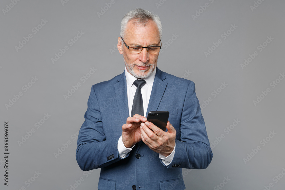 Elderly gray-haired mustache bearded business man in classic blue suit shirt tie isolated on grey background studio. Achievement career wealth business concept. Using mobile phone, typing sms message. Elderly gray-haired mustache bearded business man in classic blue suit shirt tie isolated on grey background studio. Achievement career wealth business concept. Using mobile phone, typing sms message.