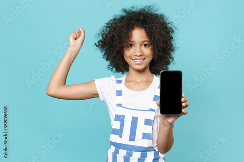 Joyful little african american kid girl 12-13 years old in striped clothes isolated on blue background. Childhood lifestyle concept. Hold mobile phone with blank empty screen doing winner gesture.