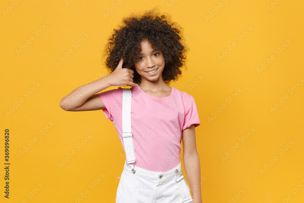 Smiling little african american kid girl 12-13 years old in pink t-shirt isolated on yellow background. Childhood lifestyle concept. Mock up copy space. Doing phone gesture like says call me back. Smiling little african american kid girl 12-13 years old in pink t-shirt isolated on yellow background. Childhood lifestyle concept. Mock up copy space. Doing phone gesture like says call me back.