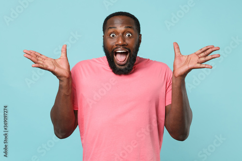 Surprised young african american man guy in casual pink t-shirt posing isolated on blue background studio portrait. People lifestyle concept. Mock up copy space. Keeping mouth open, spreading hands.