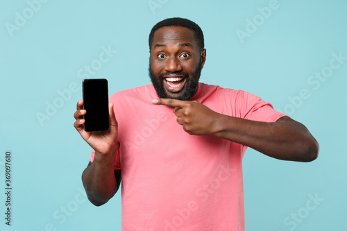 Excited young african american man guy in casual pink t-shirt isolated on blue background. People lifestyle concept. Mock up copy space. Pointing index finger on mobile phone with blank empty screen.