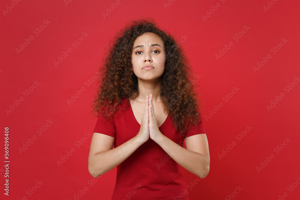 Pleading young african american woman girl in casual t-shirt posing isolated on red background studio portrait. People emotions lifestyle concept. Mock up copy space. Holding hands folded in prayer. Pleading young african american woman girl in casual t-shirt posing isolated on red background studio portrait. People emotions lifestyle concept. Mock up copy space. Holding hands folded in prayer.