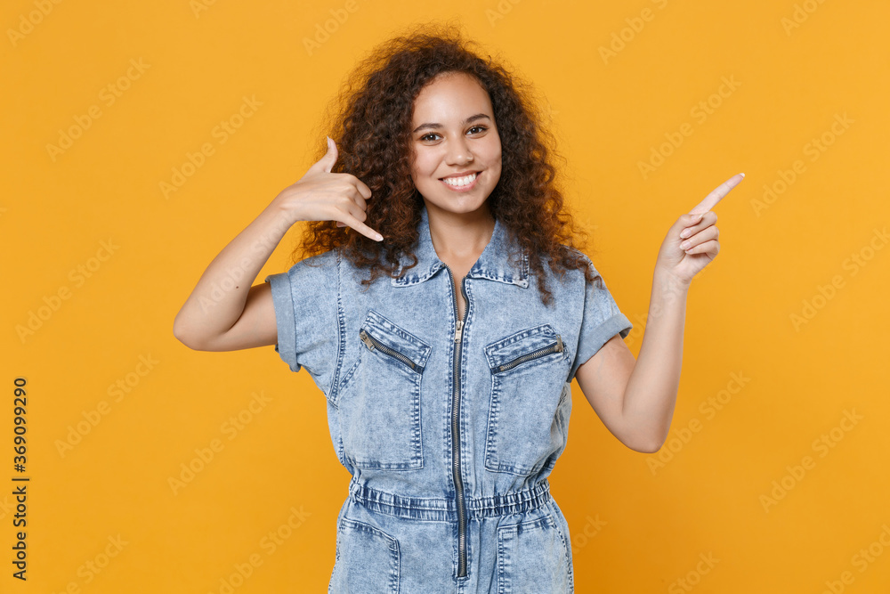 Smiling young african american woman girl in denim clothes isolated on yellow background. People lifestyle concept. Mock up copy space. Doing phone gesture says call me back, point index finger aside. Smiling young african american woman girl in denim clothes isolated on yellow background. People lifestyle concept. Mock up copy space. Doing phone gesture says call me back, point index finger aside.