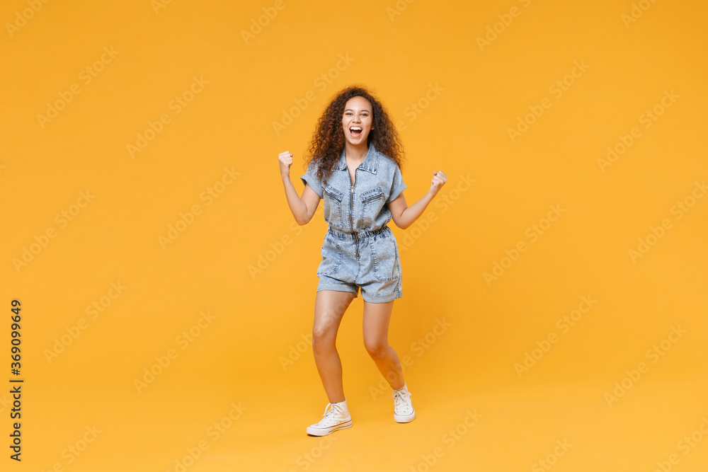 Full length portrait of joyful young african american woman girl in denim clothes isolated on yellow background studio portrait. People lifestyle concept. Mock up copy space. Doing winner gesture.