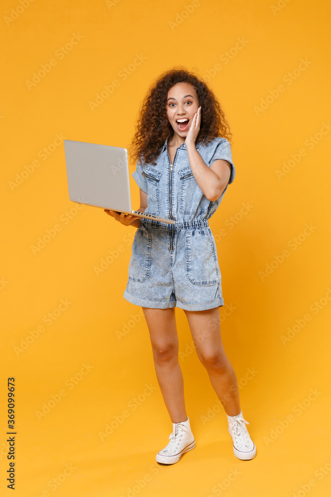Full length portrait of excited young african american woman girl in denim clothes isolated on yellow background in studio. People lifestyle concept. Working on laptop pc computer, put hand on cheek. Full length portrait of excited young african american woman girl in denim clothes isolated on yellow background in studio. People lifestyle concept. Working on laptop pc computer, put hand on cheek.