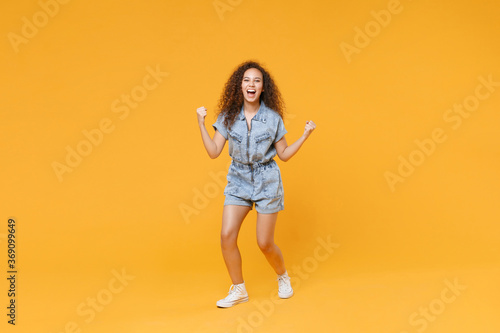 Full length portrait of joyful young african american woman girl in denim clothes isolated on yellow background studio portrait. People lifestyle concept. Mock up copy space. Doing winner gesture. © ViDi Studio