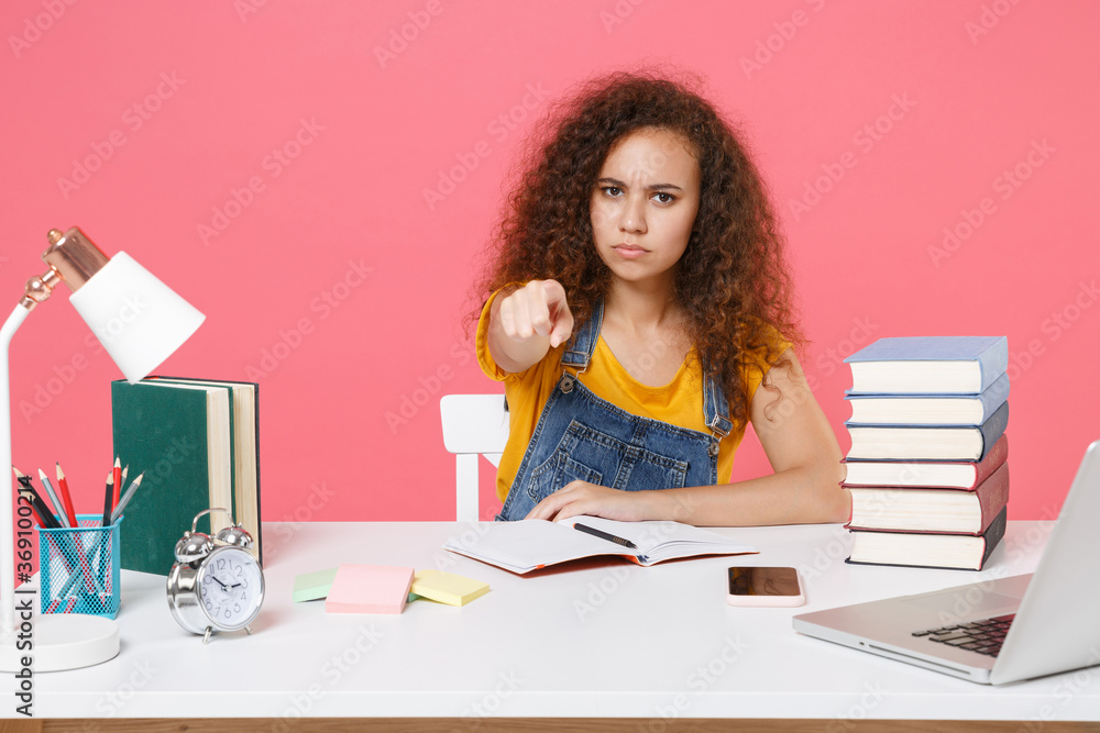 Displeased african american girl employee in office sit at desk isolated on pink background. Achievement business career. Education in school university college concept. Point index finger on camera. Displeased african american girl employee in office sit at desk isolated on pink background. Achievement business career. Education in school university college concept. Point index finger on camera.