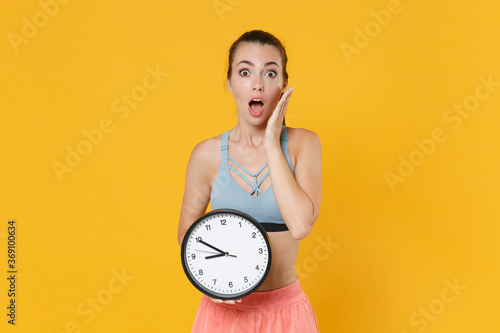 Shocked young fitness sporty woman girl in sportswear working out isolated on yellow background studio. Workout sport motivation lifestyle concept. Mock up copy space. Hold clock, put hand on cheek.