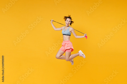 Full length portrait excited young fitness woman in sportswear working out isolated on yellow background. Workout sport motivation lifestyle concept. Mock up copy space. Jumping, hold skipping rope.