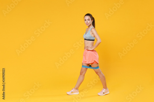 Full length portrait of young fitness sporty woman girl in sportswear working out isolated on yellow background. Workout sport motivation lifestyle concept. Doing exercise for legs with fitness gums.