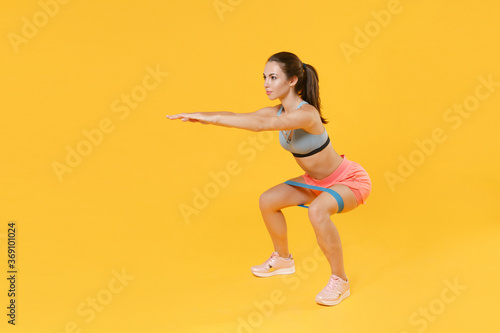 Full length portrait of young fitness sporty woman girl in sportswear working out isolated on yellow background. Workout sport motivation lifestyle concept. Doing exercise squatting with fitness gums.