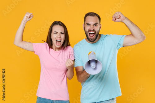 Angry young couple two friends guy girl in blue pink t-shirts posing isolated on yellow wall background studio. People lifestyle concept. Mock up copy space. Screaming in megaphone clenching fists.