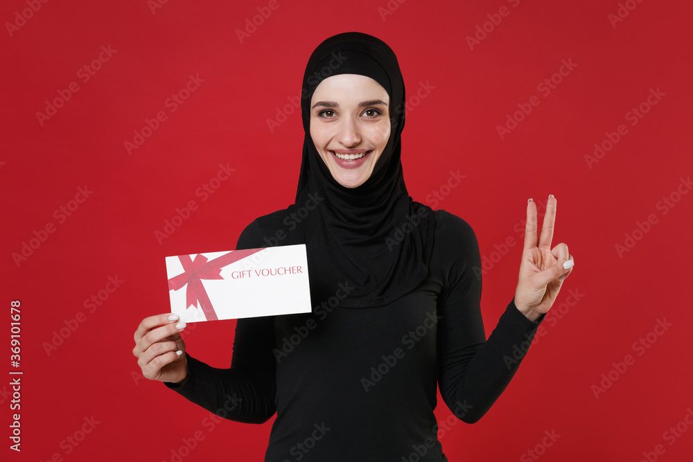 Smiling young arabian muslim woman in hijab black clothes posing isolated on bright red background. People religious lifestyle concept. Mock up copy space. Hold gift certificate, showing victory sign. Smiling young arabian muslim woman in hijab black clothes posing isolated on bright red background. People religious lifestyle concept. Mock up copy space. Hold gift certificate, showing victory sign.