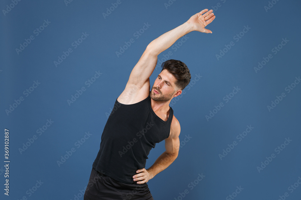Handsome young bearded fitness sporty guy sportsman in black undershirt isolated on blue wall background studio portrait. Workout sport motivation lifestyle concept. Doing stretching exercising. Handsome young bearded fitness sporty guy sportsman in black undershirt isolated on blue wall background studio portrait. Workout sport motivation lifestyle concept. Doing stretching exercising.