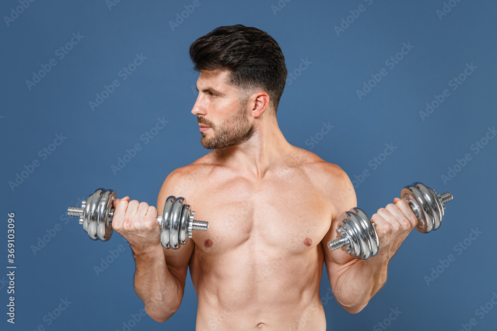 Confident young fitness sporty strong guy bare-chested muscular sportsman isolated on blue background. Workout sport motivation lifestyle concept. Doing exercise for arms with dumbbells looking aside. Confident young fitness sporty strong guy bare-chested muscular sportsman isolated on blue background. Workout sport motivation lifestyle concept. Doing exercise for arms with dumbbells looking aside.