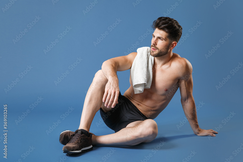 Obraz premium Full length portrait young fitness sporty strong guy bare-chested sportsman isolated on blue background. Workout sport motivation lifestyle concept. Sit on floor with towel on shoulder looking aside.