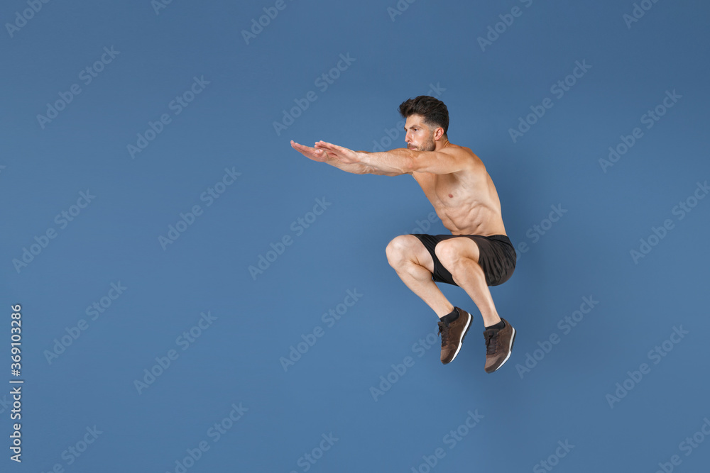 Full length portrait of young fitness sporty strong guy bare-chested muscular sportsman isolated on blue background. Workout sport motivation lifestyle concept. Jumping, doing exercises squatting. Full length portrait of young fitness sporty strong guy bare-chested muscular sportsman isolated on blue background. Workout sport motivation lifestyle concept. Jumping, doing exercises squatting.
