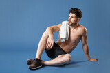 Full length portrait young fitness sporty strong guy bare-chested sportsman isolated on blue background. Workout sport motivation lifestyle concept. Sit on floor with towel on shoulder looking aside.