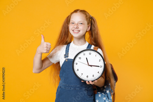 Young redhead school teen kid girl 12-13 years old in white t-shirt blue denim uniform backpack hold in hands clock isolated on yellow background children studio portrait Education lifestyle concept