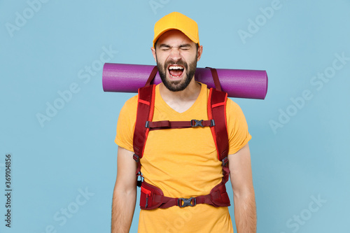 Crazy traveler young man in yellow t-shirt cap with backpack isolated on blue wall background. Tourist traveling on weekend getaway. Tourism discovering hiking concept. Keeping eyes closed screaming.