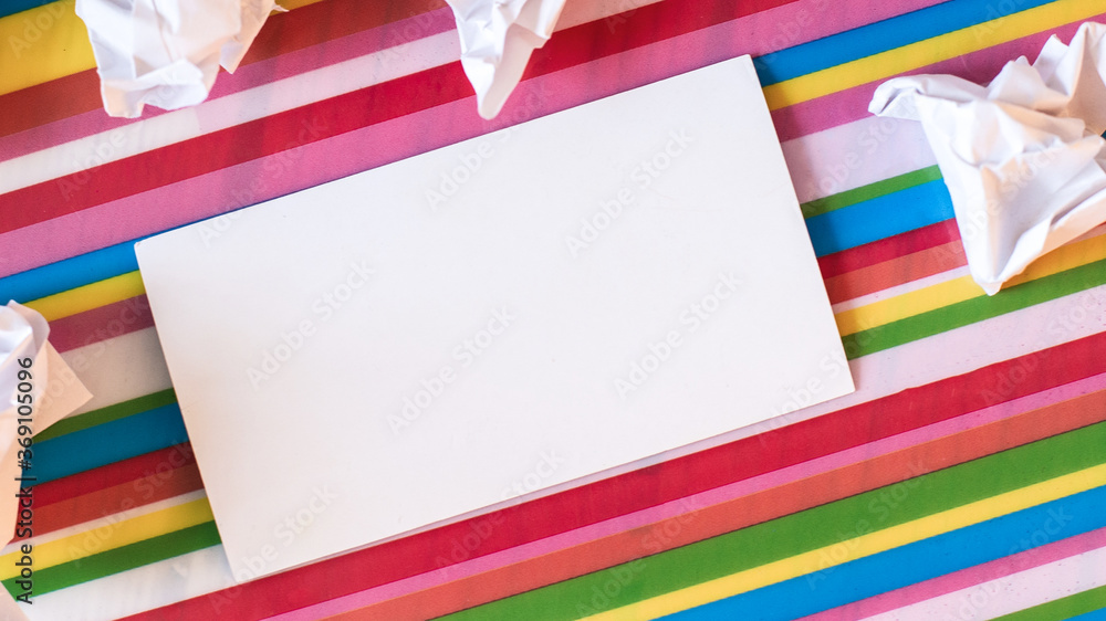 Fototapeta premium A white blank card lies on a multi-colored bright striped background near crumpled pieces of paper. Place to insert text. Close-up. Business concept