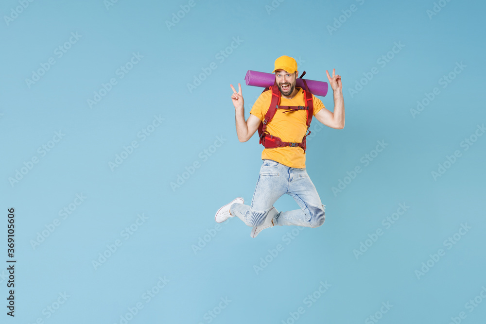 Full length portrait Excited young traveler man in cap with backpack isolated on blue background. Tourist traveling on weekend getaway. Tourism discovering hiking concept. Jump showing victory sign. Full length portrait Excited young traveler man in cap with backpack isolated on blue background. Tourist traveling on weekend getaway. Tourism discovering hiking concept. Jump showing victory sign.