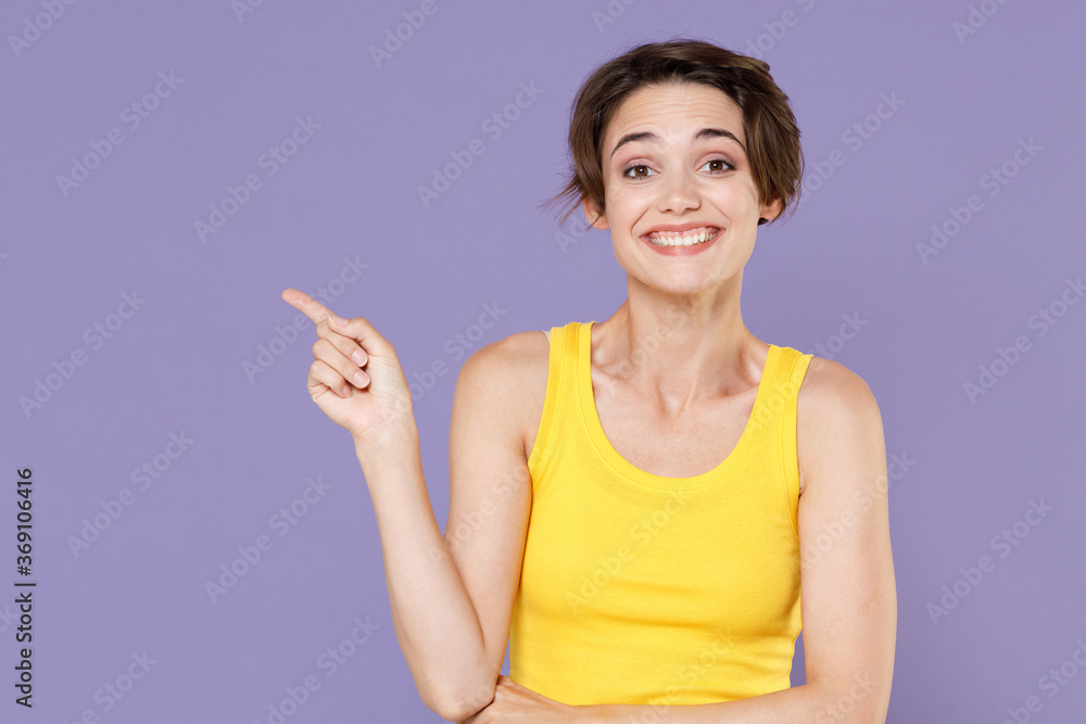 Smiling young brunette woman girl in yellow casual tank top posing isolated on pastel violet wall background studio portrait. People lifestyle concept. Mock up copy space. Pointing index finger aside. Smiling young brunette woman girl in yellow casual tank top posing isolated on pastel violet wall background studio portrait. People lifestyle concept. Mock up copy space. Pointing index finger aside.