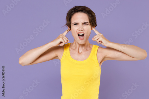 Displeased young brunette woman girl wearing yellow casual tank top posing isolated on pastel violet background studio portrait. People sincere emotions lifestyle concept. Covering ears with fingers.