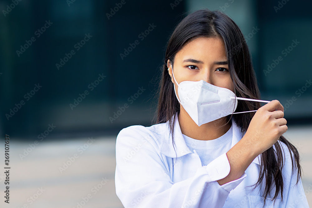 Asian woman, attractive ethnic girl in medical mask on her face looking at camera with a serious look in white shirt outside business center Asian woman, attractive ethnic girl in medical mask on her face looking at camera with a serious look in white shirt outside business center