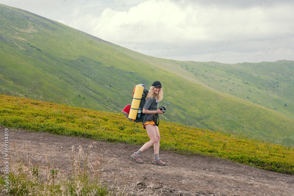 blonde girl with a backpack and a camera is walking in the mountains looking at the camera and smiling