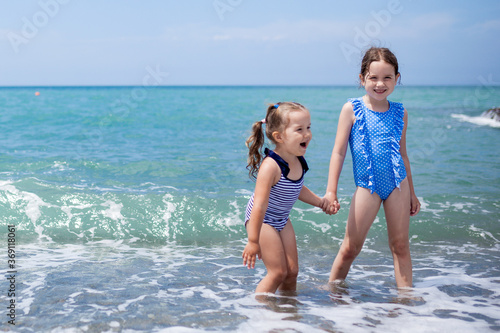 Two little girls sisters toddler having fun on the beach Children jump on the waves hold each other hands on a sunny day family vacation at the sea kids siblings leisure on the beach summer concept © tgordievskaya