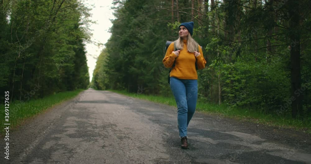 Front view, main plan: a Woman traveling on foot in a yellow sweater and hat on a forest road with a backpack and views the trees and nature. Alone on a trip. The concept of freedom and travel. Front view, main plan: a Woman traveling on foot in a yellow sweater and hat on a forest road with a backpack and views the trees and nature. Alone on a trip. The concept of freedom and travel.