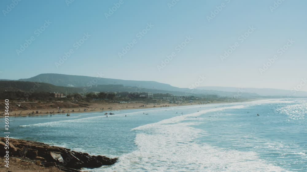 Surfing in Atlantic Ocean, Morocco, many people on the beach, big waves and clear sky and mountains on horizon, Taghazout village, full hd Surfing in Atlantic Ocean, Morocco, many people on the beach, big waves and clear sky and mountains on horizon, Taghazout village, full hd