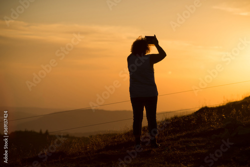Portrait  on back view of woman silhouette standing at the top of the mountain taking a photography  with his smatphone in hands on sunset background © pixarno
