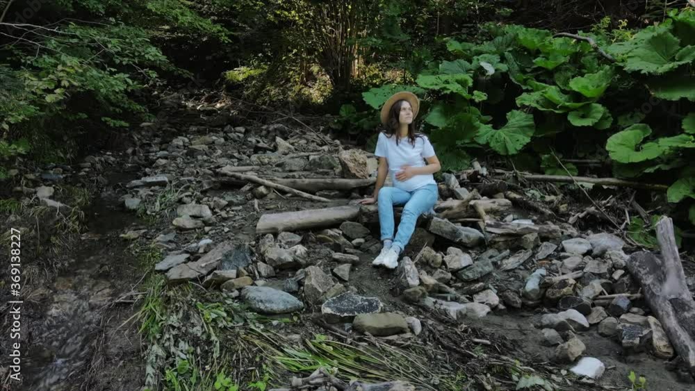 Aerial drone shot of cute young pregnant woman in hat spend time in green mixed forest near small river water, sit and relax, enjoy nature and fresh air. Concept adventure and exploring travel blog Aerial drone shot of cute young pregnant woman in hat spend time in green mixed forest near small river water, sit and relax, enjoy nature and fresh air. Concept adventure and exploring travel blog