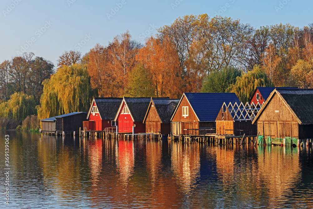 Bootshäuser an der Müritz bei Röbel in Mecklenburg-Vorpommern im Abendlicht. Mit Bäumen im Herbstlaub. Bootshäuser an der Müritz bei Röbel in Mecklenburg-Vorpommern im Abendlicht. Mit Bäumen im Herbstlaub.