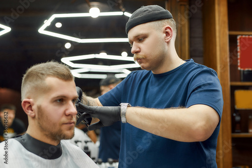 Professional barber serving young handsome man in the modern barbershop, working with hair clipper. Focus on a hairdresser