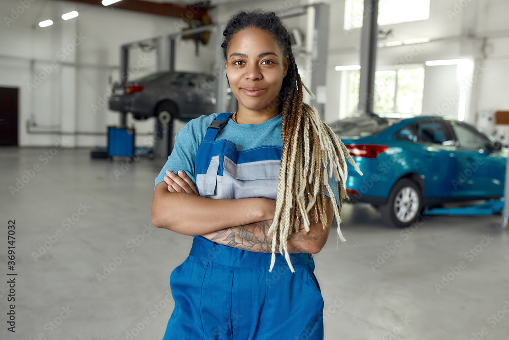 Get instant help to your car. Portrait of young african american woman,  professional female mechanic in uniform smiling at camera, standing in auto  repair shop. Car service, repair and people concept Stock-Foto