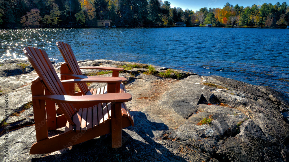 Two Muskoka chairs sitting on a rocky shore facing a calm lake with trees in the background Two Muskoka chairs sitting on a rocky shore facing a calm lake with trees in the background