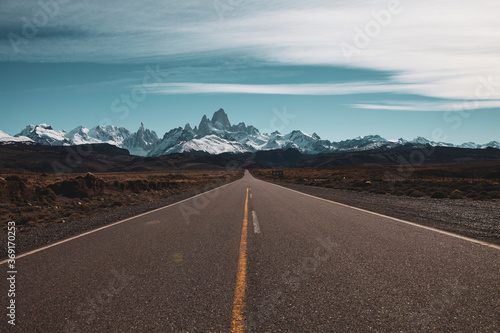 Panoramic image of a long straight asphalt road leading to frozen snowy mountains in the horizon and some clouds in the blue sky. Patagonia, Chile.