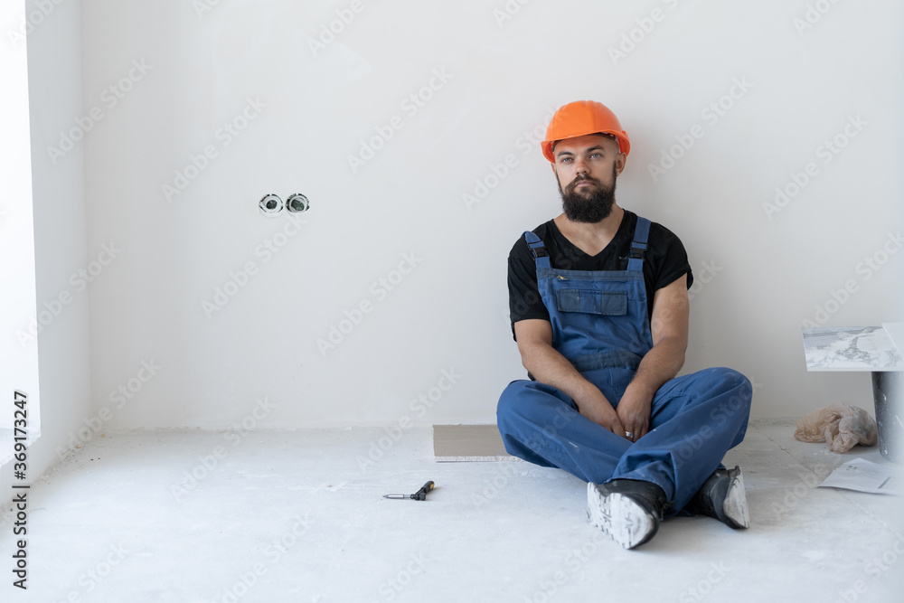 rest at the builder. a bearded man wearing a blue uniform and an orange hard hat sits on the floor during a break. Place for text. rest at the builder. a bearded man wearing a blue uniform and an orange hard hat sits on the floor during a break. Place for text.