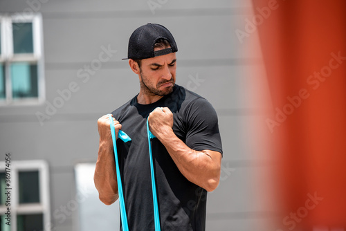 Fit muscular sports man doing bicep curl exercise with resistance band outdoors in sunlight photo