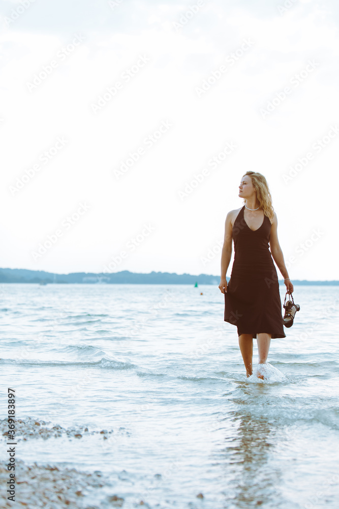 woman in black light dress walking barefoot by sea beach