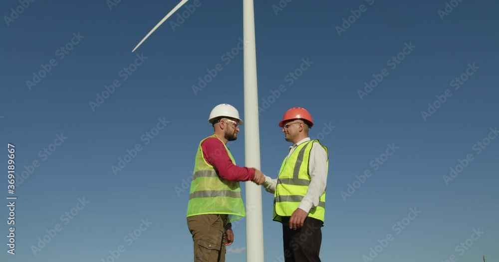 Two young engineers approach each other, shaking hands. In the background - an air windmill Two young engineers approach each other, shaking hands. In the background - an air windmill