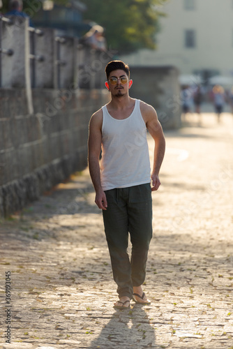 Full-length shot of young arabic man in white sleeveless shirt with sunglasses walking down a street paved with cobblestones on hot summer day looking at camera