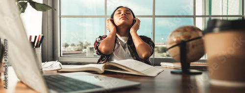 Fototapeta Naklejka Na Ścianę i Meble -  Asian young woman or female student sitting at table relax with her headphones after learning through online lessons at home. Closed eyes feels satisfaction after finish learning. Education from home.