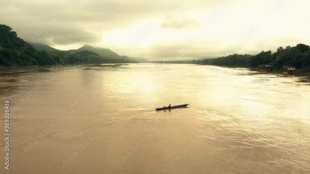 Cinematic aerial shot of local Laotian man peacefully boating down the Mekong River during sunset