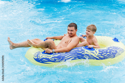 A man and a child on a yellow double rubber ring in a pool with bright blue water  close-up. Lots of spray. Two people are having fun in the water park. People ride the slides in the water park.
