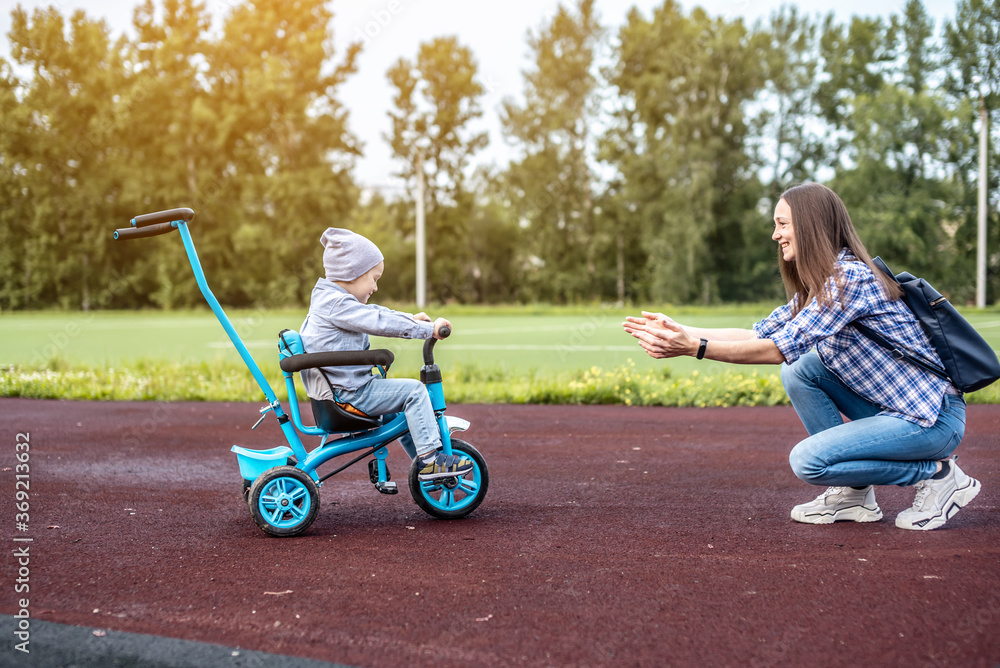 Happy toddler boy is successfully going to his mother on a children's tricycle. Concept of learning to ride a bike and having fun with your family Happy toddler boy is successfully going to his mother on a children's tricycle. Concept of learning to ride a bike and having fun with your family